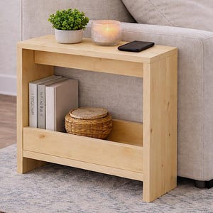 May include: A light-colored wooden side table with a shelf, holding books and a woven basket. A small potted plant, a lit candle in a glass holder, and a black smartphone sit on top. The table is next to a beige sofa.