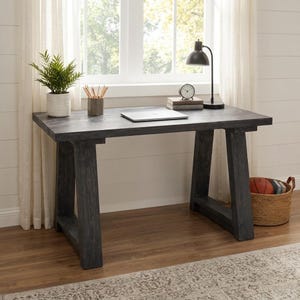 Puede incluir: Un escritorio de madera gris oscuro con una superficie rectangular y dos patas en forma de A. Sobre el escritorio hay un portátil plateado, una lámpara de mesa negra, un pequeño reloj analógico sobre dos libros y una planta verde en maceta.