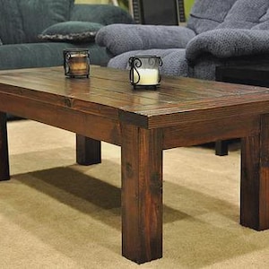 May include: A dark brown wooden coffee table with a rectangular top and four legs. The table has a distressed finish. Two candle holders with lit candles are on top. The table is in a living room setting.