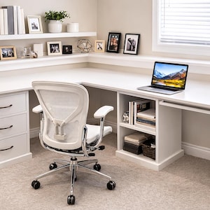 Puede incluir: Un escritorio blanco en forma de L con una silla de oficina con respaldo de malla. El escritorio tiene cajones, estantes y un ordenador portátil. Los elementos decorativos incluyen libros, plantas y fotos enmarcadas.