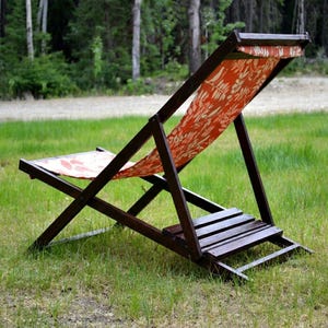 May include: A folding deck chair with a dark brown wooden frame and a fabric seat featuring an orange background with a white botanical leaf pattern. The chair has a slatted wooden footrest and a reclining backrest design for outdoor use.