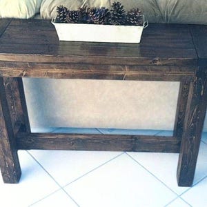 May include: A dark stained wooden side table with a rectangular top and angled legs. A white metal container filled with pinecones sits on the table's surface. The table is made of solid wood and has a rustic appearance.
