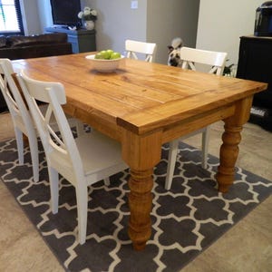 May include: A rectangular wooden dining table with a warm brown finish and thick, turned legs, accompanied by four white chairs featuring an X-back design. A white bowl containing several green apples sits in the centre of the tabletop surface.