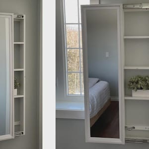 Può includere: Uno specchio rettangolare bianco montato a parete con una mensola integrata. Lo specchio è incorniciato in bianco e può essere aperto per rivelare i ripiani. I ripiani sono bianchi e contengono piante decorative. Lo specchio è montato su una parete grigio chiaro.