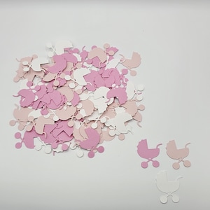 May include: A collection of paper confetti shaped like baby carriages in shades of white, light pink, and dark pink. The pieces are scattered in a pile with three individual carriage cutouts placed separately to the bottom right of the group.