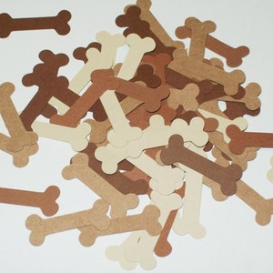May include: A pile of brown, tan, and cream paper dog bone cutouts. The bones are scattered on a white surface.