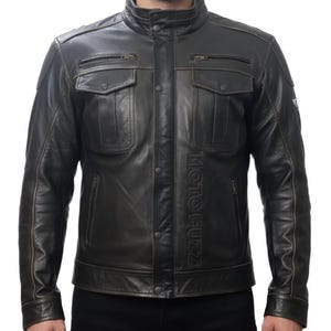 Puede incluir: Chaqueta de cuero marrón oscuro con cuello alzado, dos bolsillos en el pecho y dos bolsillos laterales. La chaqueta tiene el texto "MOTO GUZZI" verticalmente en la parte delantera. La chaqueta está abotonada y tiene un estilo clásico y robusto.
