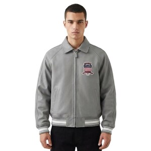 May include: A gray leather jacket with a zip-up front, a collar, and a white and gray striped hem. The jacket features a patch with a red, white, and blue design on the chest. The jacket is a classic style.