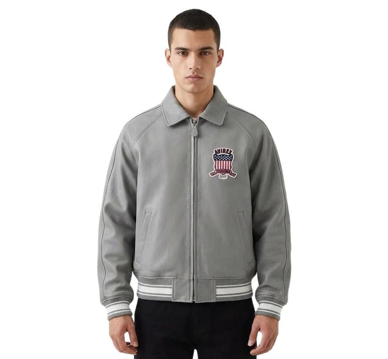 May include: A gray leather jacket with a zip-up front, a collar, and a white and gray striped hem. The jacket features a patch with a red, white, and blue design on the chest. The jacket is a classic style.