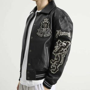 May include: Black leather jacket with white embroidered details. Features a crest and text on the chest, and a lion graphic on the sleeve. The jacket has a button-up front and a striped hem.