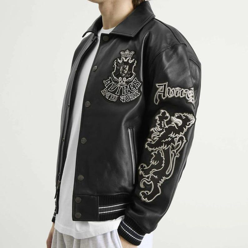 May include: Black leather jacket with white embroidered details. Features a crest and text on the chest, and a lion graphic on the sleeve. The jacket has a button-up front and a striped hem.