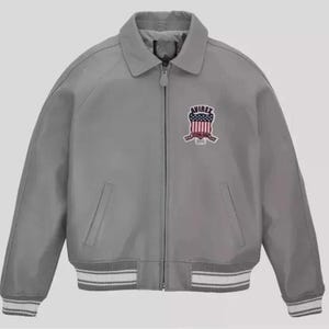 May include: A gray bomber jacket with a collar and a zipper closure. The jacket features a patch on the chest with the word "AVIREX" and a shield design in red, white, and blue. The cuffs and hem have white and gray striped detailing.