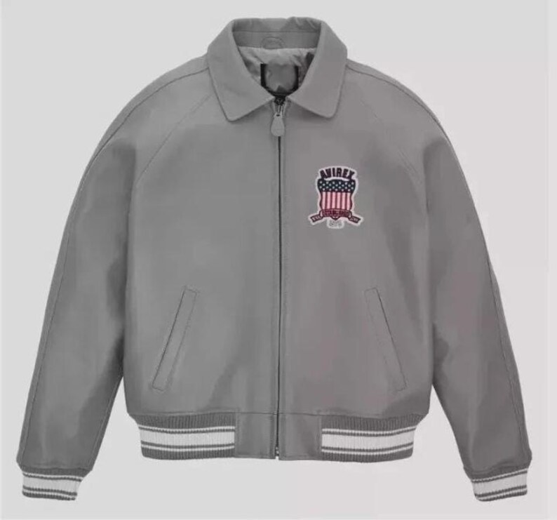 May include: A gray bomber jacket with a collar and a zipper closure. The jacket features a patch on the chest with the word "AVIREX" and a shield design in red, white, and blue. The cuffs and hem have white and gray striped detailing.