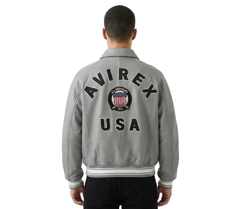 May include: A gray bomber jacket with the word "AVIREX" arched across the upper back, and "USA" below. A circular patch with red, white, and blue stripes and the words "All Americans" is centered between the text. The jacket has a collar and ribbed cuffs.