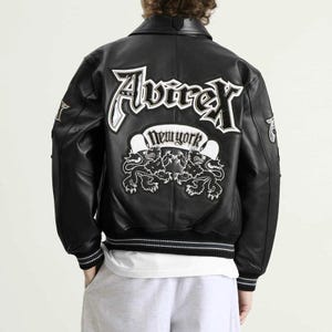 May include: Black leather jacket with white embroidered lettering and patches. The back features the word "Avirex" in large, stylized letters above a banner that reads "New York" and two lion illustrations. The jacket has a ribbed hem and cuffs.