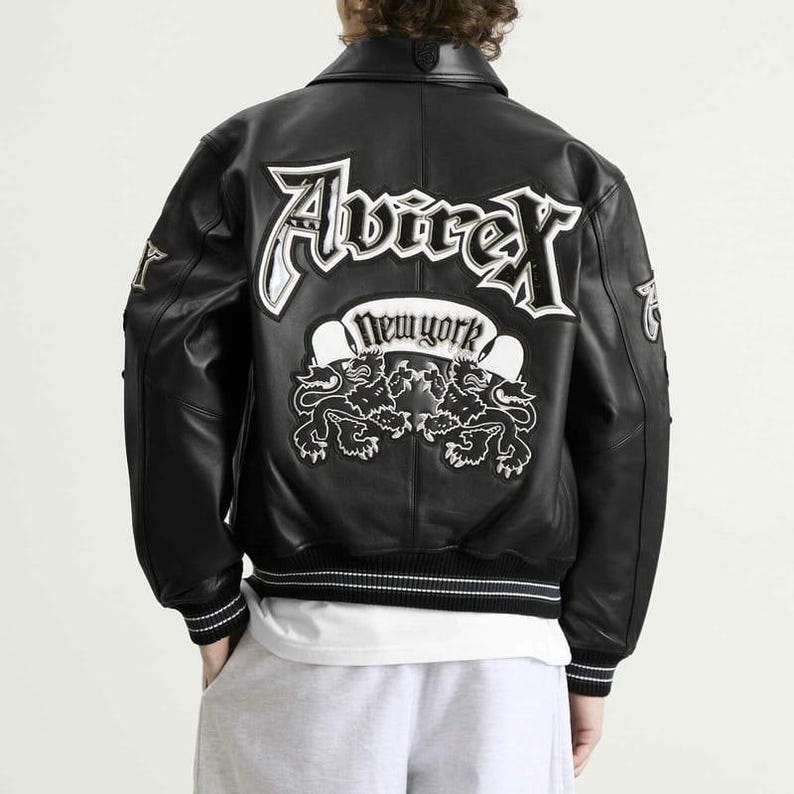 May include: Black leather jacket with white embroidered lettering and patches. The back features the word "Avirex" in large, stylized letters above a banner that reads "New York" and two lion illustrations. The jacket has a ribbed hem and cuffs.