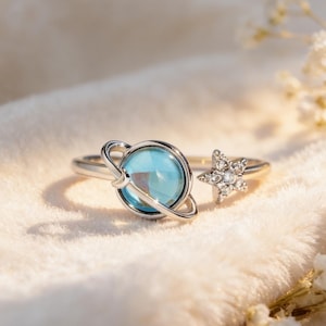 May include: A silver ring featuring a blue planet design with a band that wraps around the finger. The ring has a star-shaped accent with small, clear stones. The ring is set against a soft, white background.