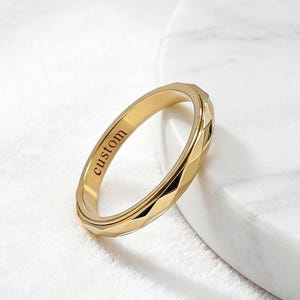 May include: A gold-toned metal ring featuring a faceted, geometric outer surface and a smooth inner band. The interior of the ring is clearly engraved with the word custom in a dark, legible serif font, showcasing the ring's polished finish.