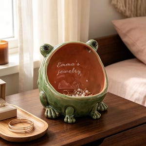 Op de afbeelding: Een groene keramische kom in de vorm van een kikker met een terracotta-kleurige binnenkant en de witte tekst "Emma's jewelry". De kom staat op een houten oppervlak naast een houten dienblad met een beige ringdoosje en goudkleurige armbanden.