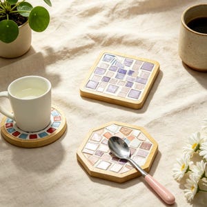 May include: Three coasters with light wood frames and mosaic tile tops. One square coaster features purple and white tiles. One round coaster holds a white mug. One octagonal coaster with pink and white tiles holds a silver spoon with a pink handle.