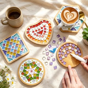 May include: A set of wooden coasters featuring mosaic designs in heart, square, and circular shapes with colorful glass tiles. One circular coaster is being decorated with purple tiles using a white adhesive tube, alongside loose glass tiles.