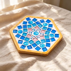 May include: A hexagonal coaster featuring a light wood frame and a mosaic surface. The mosaic consists of square and circular glass tiles in shades of blue, light blue, and soft pink, arranged in a geometric pattern on a white base.