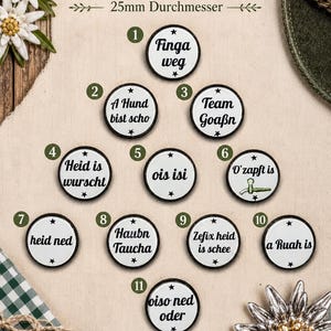 May include: Eleven round white pins with black text and borders, each 25mm in diameter. The pins feature various German phrases including Finga weg, A Hund bist scho, Team Goaßn, Heid is wurscht, ois isi, O'zapft is, heid ned, Haubn Taucha, Zefix heid is schee, a Ruah is, and oiso ned oder.