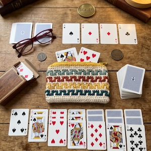 May include: A small, rectangular, crocheted zipper pouch featuring a geometric pattern in cream, yellow, teal, and rust-orange yarn. The pouch is surrounded by various playing cards, a wooden card box, red-rimmed glasses, and metal dealer coins.