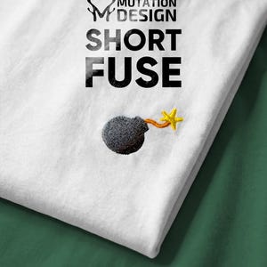 May include: White t-shirt with the text "MUTATION DESIGN SHORT FUSE" in black. A small embroidered bomb with a yellow star-shaped fuse is on the lower left. The shirt is folded on a green surface.