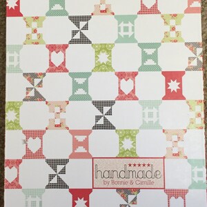 May include: A white box featuring a repeating pattern of colourful spools, hearts, and stars. The spools are in shades of red, green, and black. The box has a decorative border and the text "handmade by Bonnie & Camille".