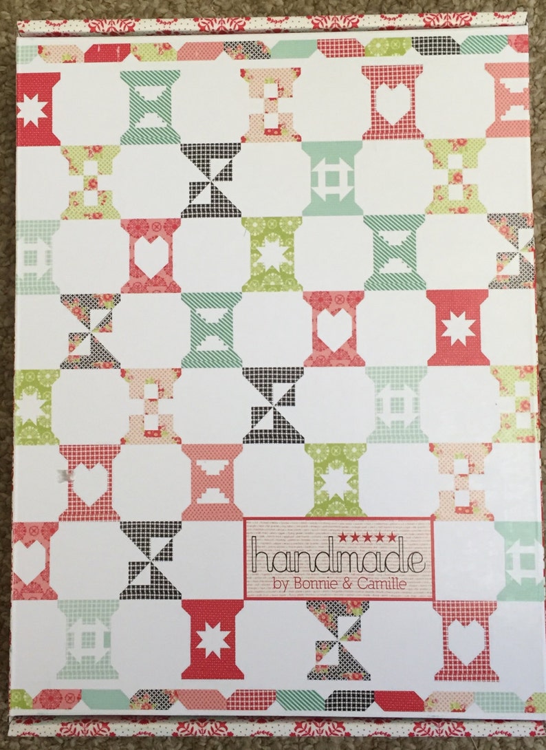 May include: A white box featuring a repeating pattern of colourful spools, hearts, and stars. The spools are in shades of red, green, and black. The box has a decorative border and the text "handmade by Bonnie & Camille".