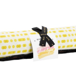 May include: A rolled fabric bundle with a white background and yellow dot pattern. The bundle is tied with a black ribbon and a tag that reads "first light". The fabric edges are black.