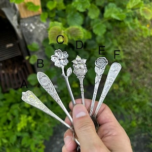 Puede incluir: Una colección de seis cucharas de metal plateado etiquetadas de la A a la F, cada una con distintos diseños florales o botánicos en relieve en los mangos. Las cucharas están dispuestas en abanico, mostrando patrones intrincados de rosas, margaritas y otras flores.