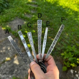 Könnte beinhalten: Sechs silberfarbene Metalllöffel mit unterschiedlichen dekorativen Mustern auf den Griffen, beschriftet von A bis F. Jeder Griff weist ein einzigartiges geprägtes Design auf, darunter florale, geometrische und lineare Motive, fächerförmig angeordnet.