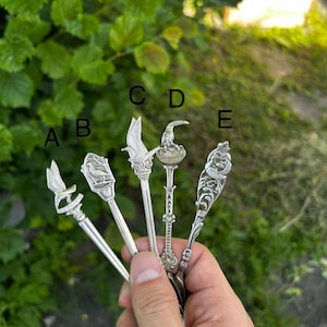 May include: Five silver-toned metal souvenir spoons labeled A, B, C, D, and E. Each spoon features a unique decorative handle top, including bird motifs and intricate filigree patterns. The spoons are held together in a fan shape.