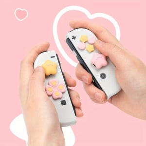 Peut inclure: Manettes de jeu blanches et noires avec des couvre-boutons colorés en forme d'étoile et de fleur. Les manettes sont tenues sur un fond rose avec un contour de cœur blanc.