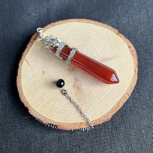 Puede incluir: Un péndulo de cristal de cornalina roja con una corona y cadena de metal plateado, y una cuenta negra. El péndulo se muestra sobre una rodaja de madera.