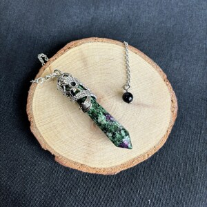 May include: A green and purple crystal pendulum with a silver-colored metal cap and chain, and a black bead at the end of the chain. The pendulum is resting on a round wooden slice.