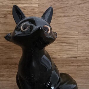 Könnte beinhalten: Glänzende schwarze Fuchsskulptur mit spitzen Ohren und einem eingerollten Schwanz. Der Fuchs sitzt, mit detaillierten Gesichtszügen und hellbraunen Augen. Die Figur ist ein dekoratives Objekt.