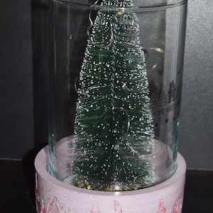 May include: A decorative Christmas tree inside a clear glass cylinder. The tree is green with white accents and fairy lights. The cylinder sits on a pink base with a gnome design. The words "HOME SWEET HOME" are visible on the glass.