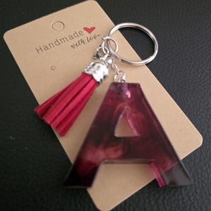 Könnte beinhalten: Ein burgunderfarbener Schlüsselanhänger mit dem Buchstaben "A", einem passenden Quastenanhänger und silberfarbener Hardware. Der Schlüsselanhänger ist an einem braunen Anhänger mit der Aufschrift "Handmade with love" befestigt.