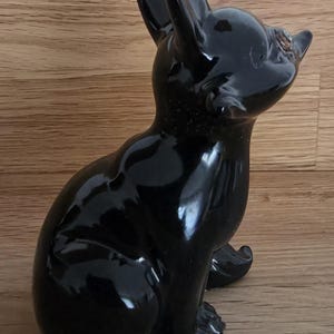 May include: A glossy black ceramic dog figurine in a seated position, with upright ears and a focused gaze. The figurine has a smooth, reflective surface and is set against a wooden background.