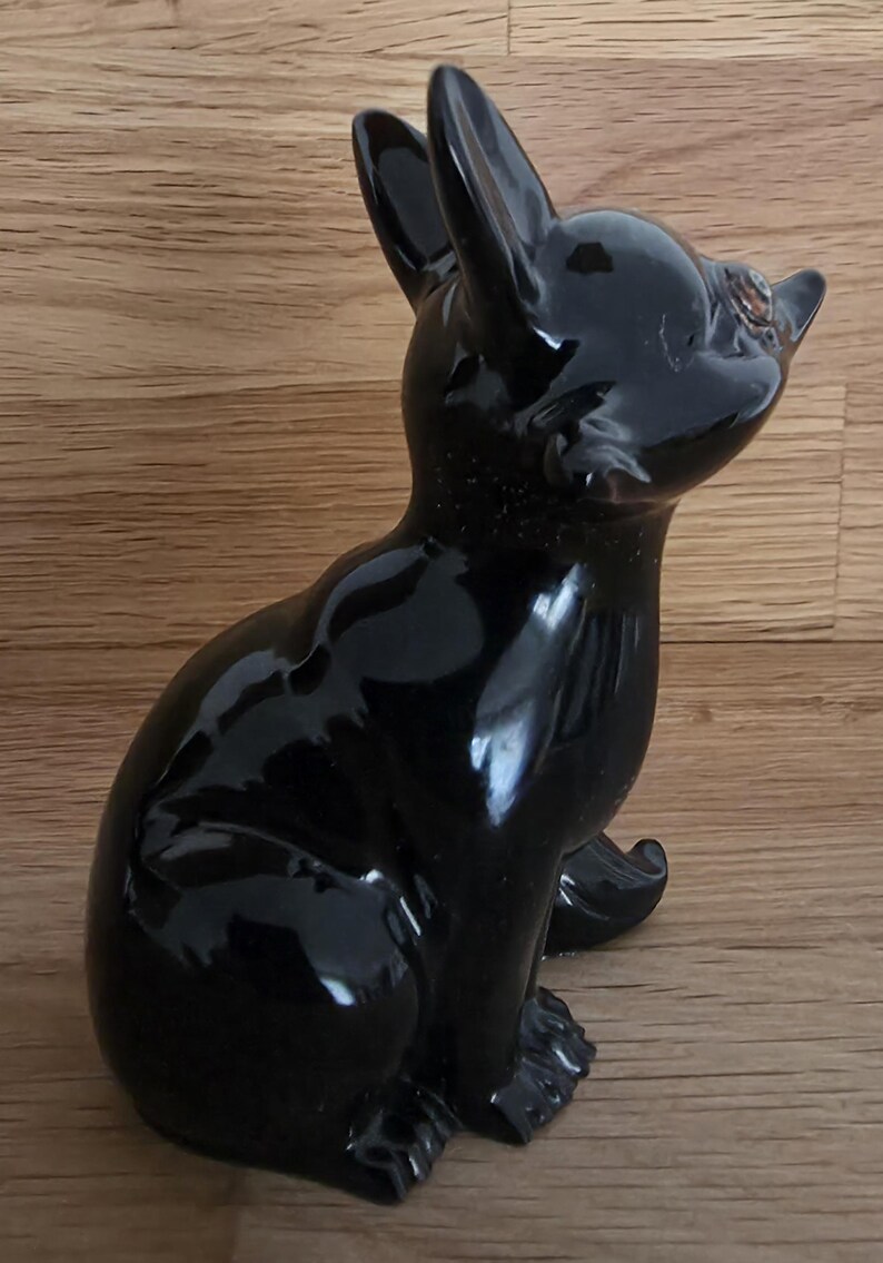 May include: A glossy black ceramic dog figurine in a seated position, with upright ears and a focused gaze. The figurine has a smooth, reflective surface and is set against a wooden background.