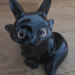May include: A glossy black ceramic figurine of a stylized animal, possibly a cat or fox, with large ears and golden eyes. The figurine has a sleek, polished finish and is posed in a seated position. The figurine is on a wooden surface.