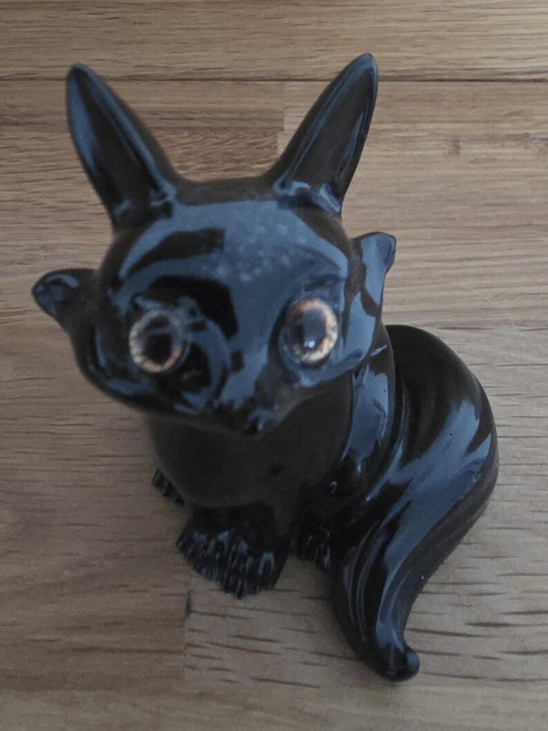 May include: A glossy black ceramic figurine of a stylized animal, possibly a cat or fox, with large ears and golden eyes. The figurine has a sleek, polished finish and is posed in a seated position. The figurine is on a wooden surface.