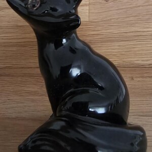 May include: A glossy black ceramic fox figurine. The fox is sitting with its head turned to the left, ears perked up, and a long tail curled around its body. The figurine has detailed features and a reflective surface.