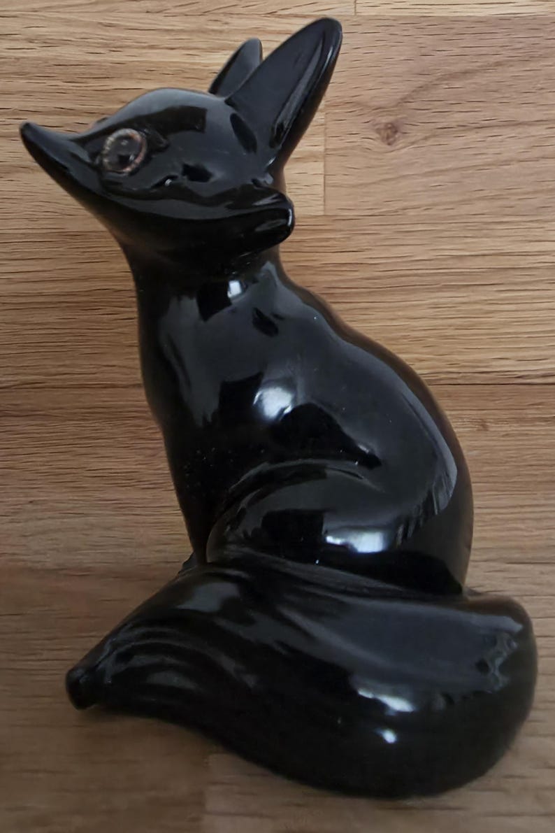 May include: A glossy black ceramic fox figurine. The fox is sitting with its head turned to the left, ears perked up, and a long tail curled around its body. The figurine has detailed features and a reflective surface.