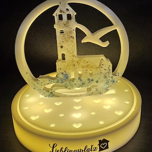 May include: A decorative night light featuring a lighthouse and seagull design within a circular frame. The light sits atop a base with heart-shaped cutouts and the text "Lieblingsplatz". The light is yellow and has a glittery effect.
