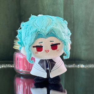 Puede incluir: Muñeco de peluche con pelo turquesa y ojos rojos, vistiendo un abrigo blanco con detalles negros. El muñeco tiene una pequeña cara sonriente y zapatos negros. Un tarro rosa es visible en el fondo.