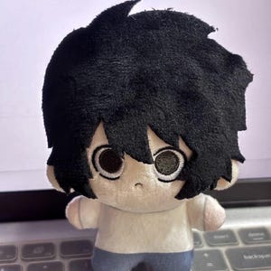 May include: A plush doll of a character with black, fluffy hair and large, round eyes. The doll wears a white shirt and blue pants. The doll is a collectible item.